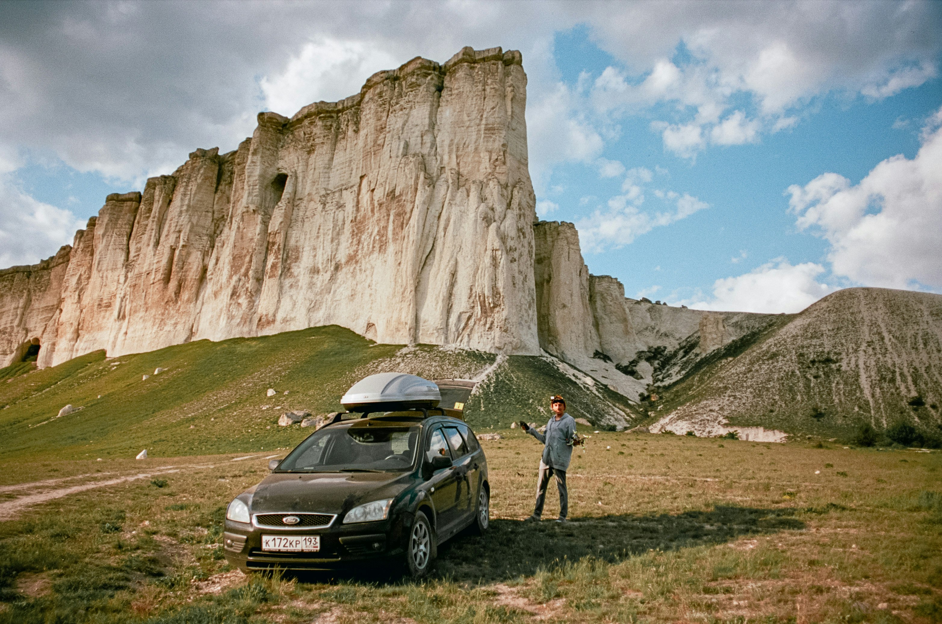 man in gray shirt and blue denim jeans standing beside black car near brown rocky mountain